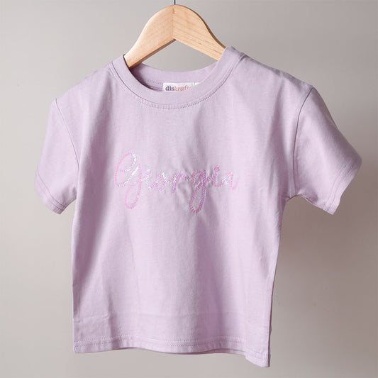 Lilac drop shoulder tee with 'Giorgia' embellished in iridescent deep pink sequin hanging on a wooden hanger against a plain background.